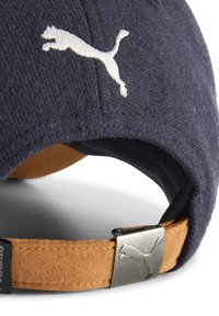 Gorra azul marino hecha de tela texturizada, con un logo blanco de Puma y una correa de ante marrón con un detalle metálico de Puma para ajuste.