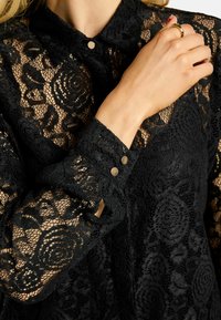 Melna mežģīņu blūze ar apkaklīti, sarežģītiem ziedu rakstiem un pogām piedurknēs. Caurspīdīga tekstūra ar detalizētu izšuvumu.