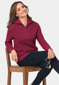 Weinroter Pullover mit einem hohen Kragen und halbem Reißverschluss, strukturiertes Strickmuster, kombiniert mit marineblauen Skinny-Jeans. Holzstuhl mit Armlehne.