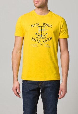 Gelbes Baumwoll-T-Shirt mit rundem Ausschnitt. Mit navyblauem Textdruck und einem Anker-Motiv mittig auf der Brust. Kurze Ärmel.