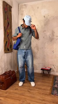 Άνδρας με πουκάμισο καρό, μπλε τζιν, λευκά αθλητικά παπούτσια και ανοιχτό μπλε καπέλο, κρατώντας μια μπλε τσάντα σε ένα ρουστίκ δωμάτιο με καφέ δερμάτινη τσάντα και ξύλινο σκαμπό.