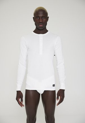 Langærmet hvid henley skjorte lavet af ribbet bomuld, med en knaplukning og et mærke i kanten. Glat tekstur med tætsiddende design.
