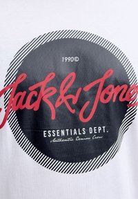 Valge T-särk, millel on ringikujuline graafiline disain. Must ja punane tekst ütleb "Jack & Jones", koos "Essentials Dept." sildi ja triibuliste aktsentidega.