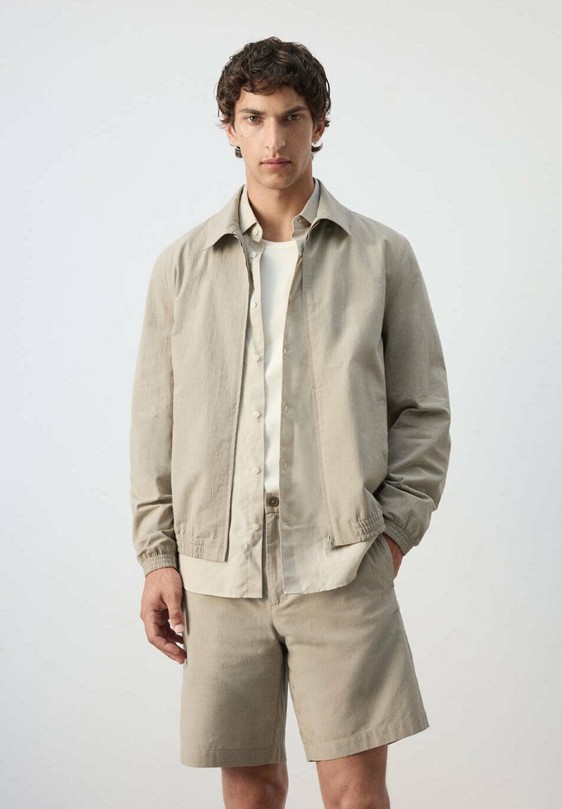 Jeune homme portant une veste beige clair, un short assorti et une chemise blanche, se tenant devant un fond clair uni.