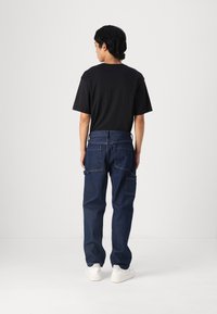 Pantalones cargo de denim azul oscuro con múltiples bolsillos, combinados con una camiseta negra lisa y zapatillas blancas, vistos desde atrás.