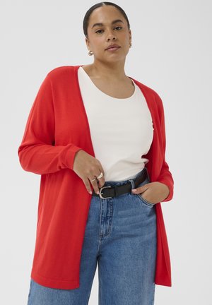 Žena v červenom kardigáne cez biely top a modré džínsy, upravujúca čierny opasok, stojaca pred jednofarebným svetlosivým pozadím.