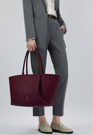 Vrouw in grijs pak met een grote bordeauxleren tote bag, gekleed in beige loafers, staande tegen een lichtgrijze achtergrond.