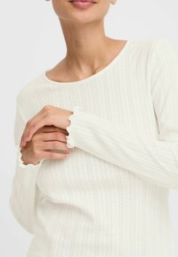 Witte lange mouw top met een geribde textuur, voorzien van een ronde halslijn en gekartelde manchetten. Subtiele verticale strepen en kleine patronen.