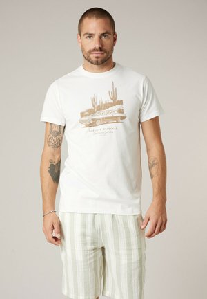 Homme avec barbe et cheveux courts portant un t-shirt blanc avec un motif de voiture et de cactus, short rayé blanc et vert, debout devant un fond uni.