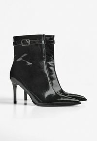 Ghete de gleznă din piele lacuită neagră, cu vârf ascuțit, toc înalt și cataramă reglabilă la gleznă. Textură netedă și lucioasă.