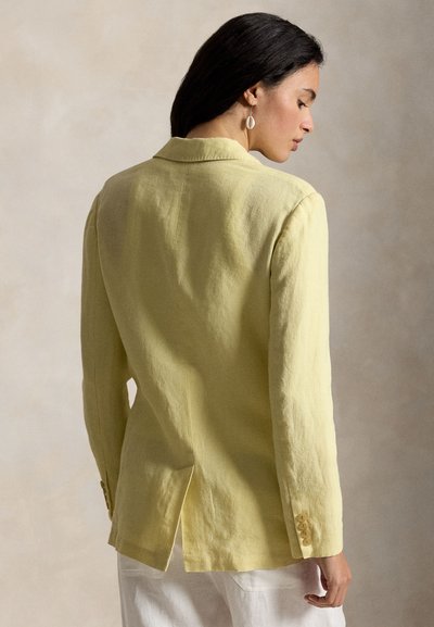 Femme aux cheveux foncés portant un blazer jaune clair et un pantalon blanc, vue de profil sur un fond neutre uni.