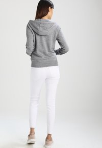 Šedá mikina s kapucí a dlouhými rukávy, s žebrovanými manžetami a lemem. Nošena s bílými džínami a světlými teniskami. Pohled zezadu.