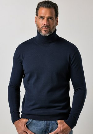 Mann mit kurzen dunklen Haaren und Bart, der einen marineblauen Rollkragenpullover und blaue Jeans trägt, mit den Händen in den Taschen vor einem einfarbigen Hintergrund.