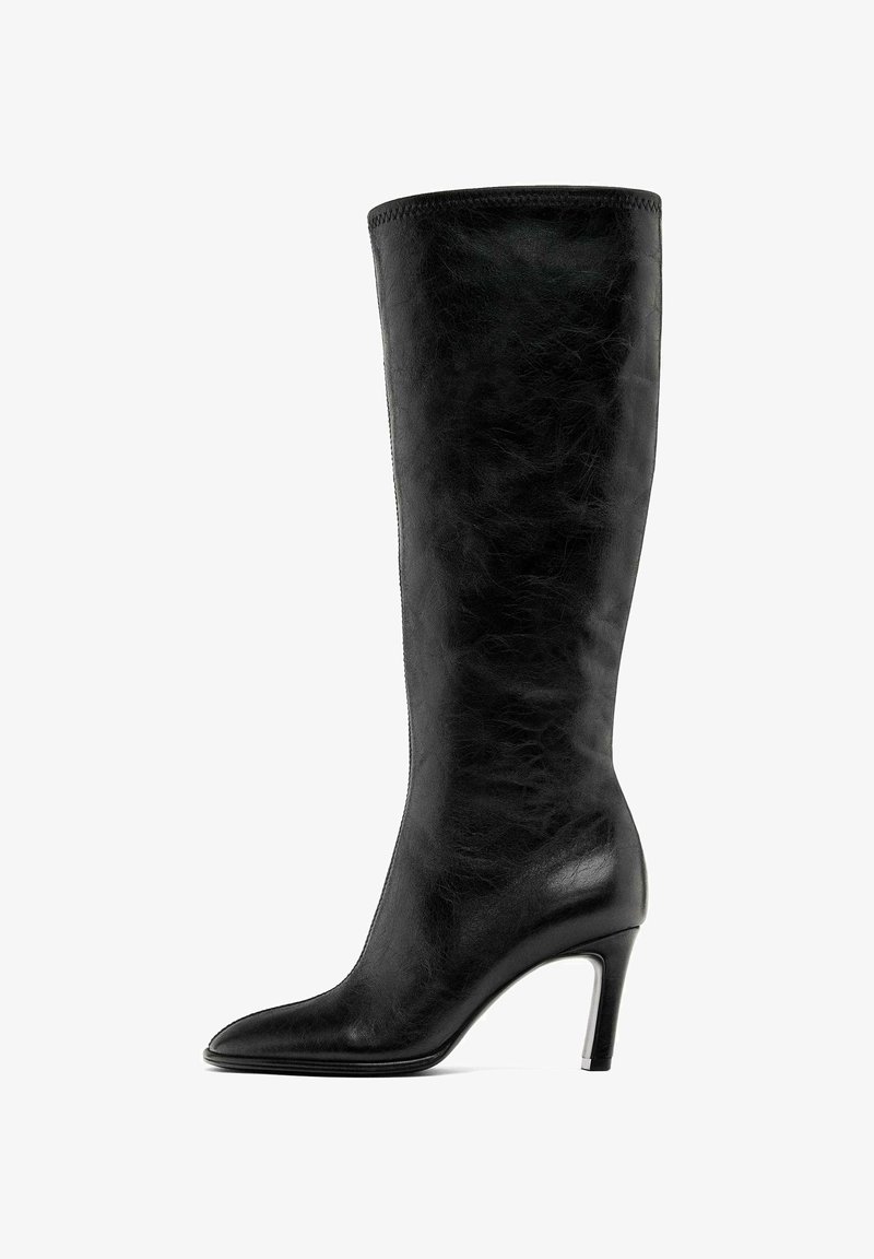 Botte noir en cuir, hauteur genou, avec un bout pointu et un talon aigu slim. Texture lisse avec un minimum de couture et un design épuré.