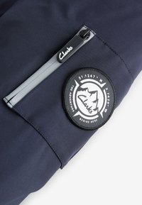 Poche zippée sur tissu bleu marine avec curseur de fermeture éclair avec le logo Clarks et écusson circulaire représentant des montagnes et des coordonnées.