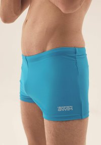 Niebieskie kąpielówki o gładkiej strukturze, z białym logo na dolnej lewej stronie. Projekt jest dopasowany i ma długość do połowy uda.