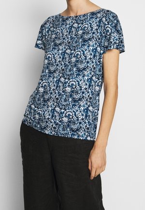 Femme portant un chemisier à manches courtes bleu et blanc à motif floral, associé à un pantalon noir ample, sur un fond uni.