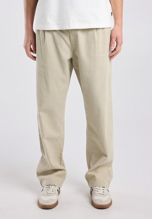 Mann trägt lockere beige Hosen, weiße Sneakers und ein weißes Hemd, steht vor einem schlichten hellgrauen Hintergrund.