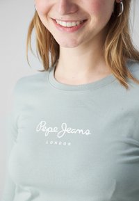 Šviesiai žalia ilgomis rankovėmis marškinėliai su balta "Pepe Jeans" logotipo spauda. Lygus audinys, apvalios iškirptės ir minimalistiškas dizainas.