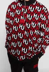 Sudadera negra con cremallera que presenta un patrón repetido de letras rojas y blancas "GO" y "HU", con una pequeña etiqueta roja que muestra "HUGO".