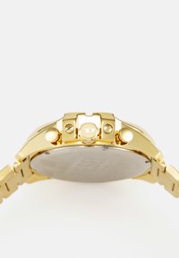 Orologio in metallo dorato con cassa rotonda, lunetta testurizzata e cinturino liscio a contrasto. Presenta una corona e pulsanti prominenti. Design minimalista.