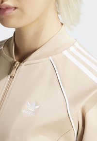 Chaqueta beige con cremallera y detalles en blanco con tres franjas. Presenta el logotipo de adidas bordado en blanco en el lado izquierdo del pecho y un cuello de punto.