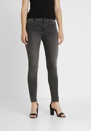 Kvinde iført sort ærmeløs top, mørkegrå skinny jeans og sorte åbne højhælede sandaler, stående på hvid baggrund.