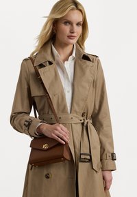 Brun läderhandväska med en guldsymbol, med lockstängning och en axelrem, hålls av en person i en beige trenchcoat över en vit skjorta.