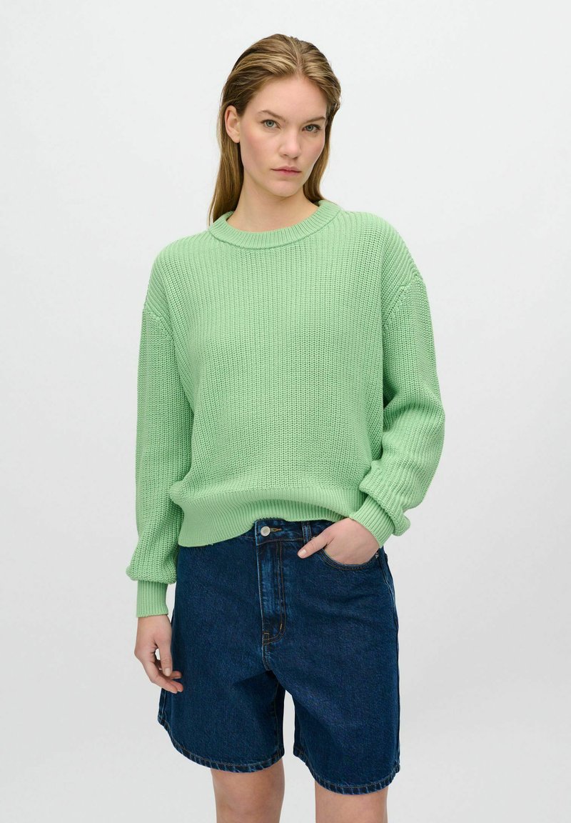 Hellgrüner Strickpullover mit Rundhalsausschnitt, überschnittenen Schultern und elastischen Bündchen, kombiniert mit dunkelblauen Jeansshorts.