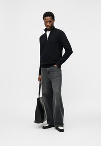 Άνδρας που φορά μαύρο πουλόβερ με φερμουάρ μέχρι το στήθος, φαρδιά γκρι τζιν παντελόνα, ασπρόμαυρα παπούτσια, κρατώντας μαύρη τσάντα ώμου, στέκεται με το ένα χέρι στην τσέπη.