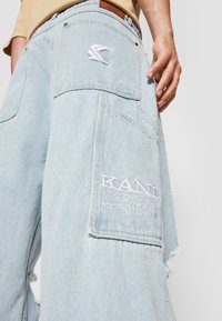 Pantalones cargo de mezclilla azul claro con parches bordados. Presentan múltiples bolsillos, dobladillos deshilachados y una etiqueta de cuero en la cintura.