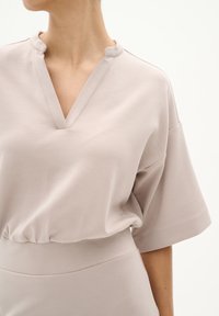 Helles beige Bluse mit V-Ausschnitt und einem dezenten Kragen. Verfügt über kurze, weite Ärmel und einen gerafften Saum für eine lässige Passform. Glatte Textur.