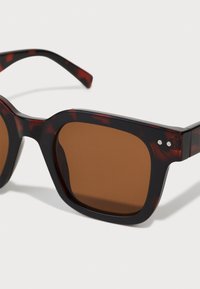 Lunettes de soleil teintées marron avec une monture en acétate à motif écaille de tortue épaisse. Le design présente une forme carrée et des accents métalliques sur les branches.