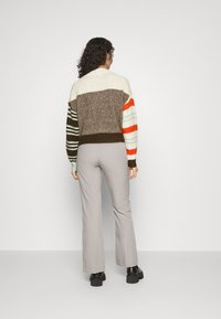 Strikket sweater med multikolor striber i brun, creme og orange; ribbestrikket kant og manchetter. Parret med lysegrå brede bukser og sorte sko.
