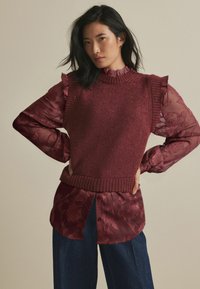 Pull en tricot bordeaux avec col côtelé superposé sur une chemise en voile à motifs floraux. Le mannequin porte un pantalon en denim ample.