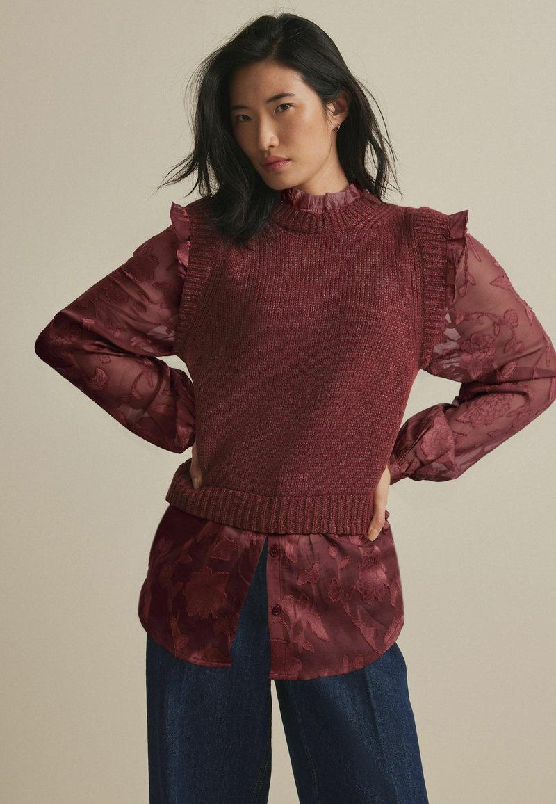 Pull en tricot bordeaux avec col côtelé superposé sur une chemise en voile à motifs floraux. Le mannequin porte un pantalon en denim ample.