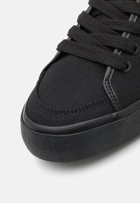 Baskets en toile noire à surface texturée, bout rond et semelle plate en caoutchouc noir. Présente des lacets et œillets noirs. Design minimaliste.