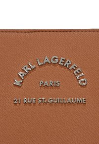 Hnedá textúrovaná koža s vyvýšeným strieborným textom: "KARL LAGERFELD," "PARÍŽ," a "21 RUE ST. GUILLAUME" v zakrivenom usporiadaní.