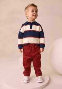 Gestreiftes Polo in Marineblau und Creme mit roten Akzenten, kombiniert mit weiten roten Cordhosen; ein Kind in weißen Turnschuhen steht auf einer runden Plattform.
