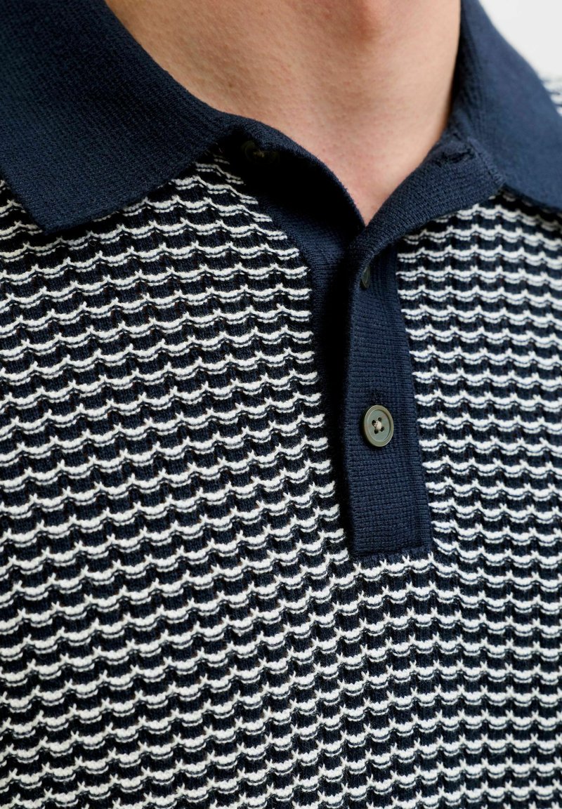 Gros plan sur un polo en maille texturée bleu marine et blanche avec un col foncé et deux boutons noirs, porté sur la partie supérieure de la poitrine d'une personne.