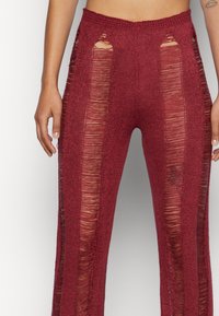 Detailní záběr na osobu, která má na sobě červené pletené kalhoty s vysokým pasem, které mají podélné průsvitné třásňové panely po přední části a po stranách.