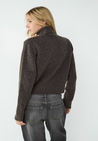 Dunkelbrauner, eng geschnittener Pullover mit hohem Kragen und gerippten Bündchen, kombiniert mit hoch taillierten, verblichenen schwarzen Jeans, von hinten gezeigt.