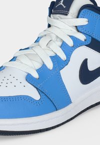 Μπλε και λευκό sneaker υψηλού τύπου με λευκά κορδόνια, διατρητό κουτί δαχτύλων, μαύρο λογότυπο Nike Swoosh και λογότυπο Jordan στη γλώσσα.