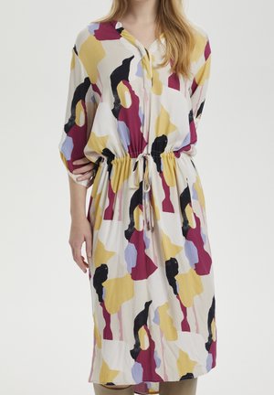 Kvinde iført en langærmet kjole med et abstrakt mønster i gult, sort, bordeaux og lavendel samt en justerbar snor i taljen.