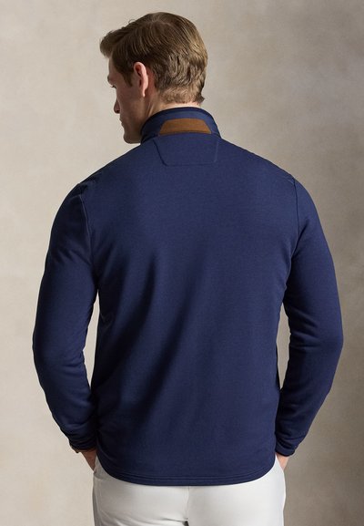 Hombre con cabello castaño claro y corto, vistiendo una camisa de manga larga azul marino con un parche marrón en el cuello trasero, sobre un fondo neutro.