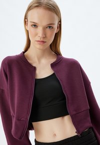 Chaqueta morada recortada con cremallera y puños acanalados, combinada con una camiseta negra ajustada, que muestra un diseño deportivo y casual.