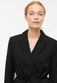 Blazer nero su misura con collo a revers e design avvolgente. Presenta una cintura annodata in vita e un tessuto leggermente strutturato.