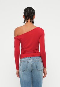 Czerwona bluzka z długim rękawem o asymetrycznym dekolcie, z dopasowanym krojem i marszczonym materiałem w talii, w połączeniu z niebieskimi dżinsami.