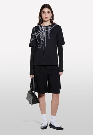 T-shirt noir avec un design en chaîne argentée ; associé à un short noir. Le modèle tient un sac à main noir, portant des chaussettes blanches et des ballerines noires.