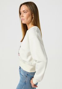 Sudadera blanca de cuello redondo y mangas largas, con un diseño gráfico en el frente, combinada con jeans de mezclilla azul. Textura de tela suave.